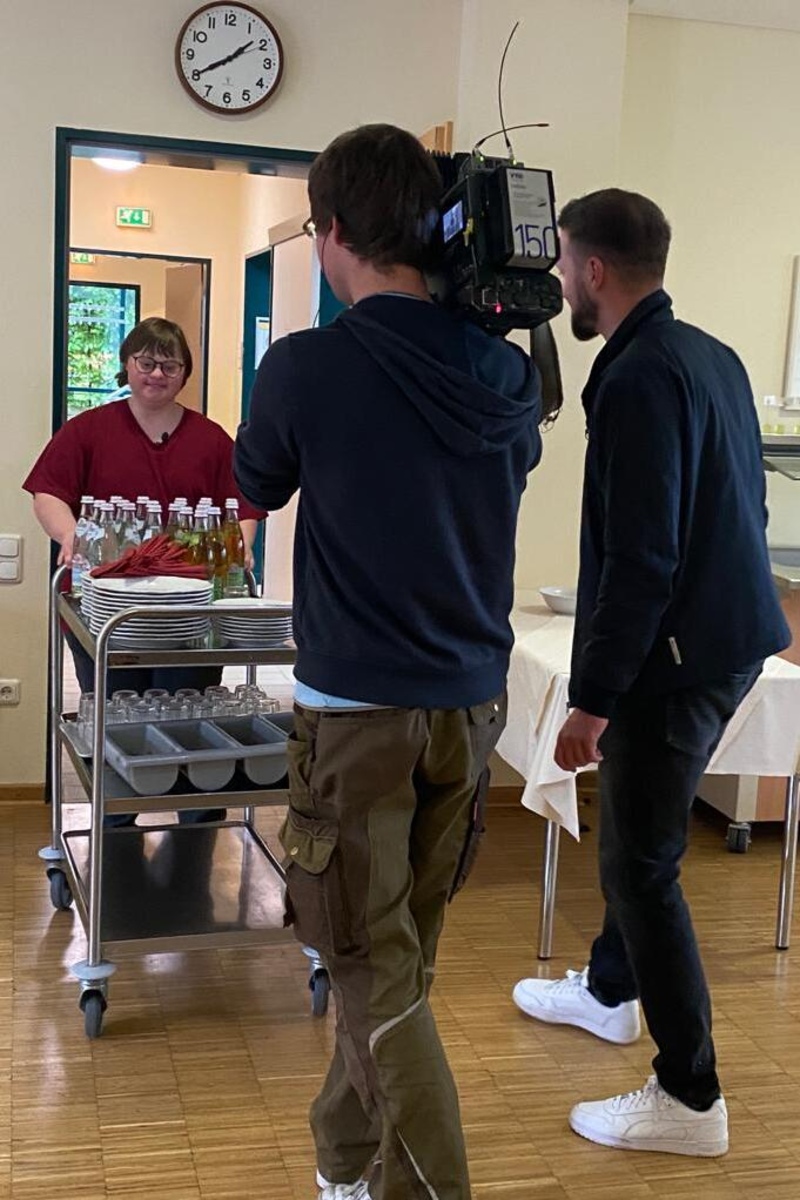 Mitarbeiterin schiebt Servierwagen. Ein Filmteam filmt sie dabei.