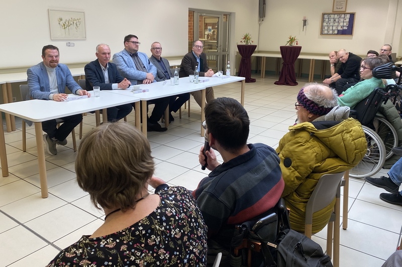 Politiker sitzen vor den Gästen der Podiumsdiskussion. Ein Mitarbeiter der KJF Werkstätten stellt ihnen eine Frage.
