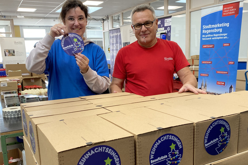Eine Mitarbeiterin und ein Mitarbeiter der KJF Werkstätten stehen hinter einem Stapel fertiger Päckchen. Sie freuen sich über die Aktion mit Hannes Ringlstetter.
