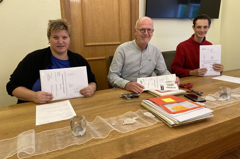 Ken-Roy Hummel (rechts) und Verena Klein (links) halten Dokumente in die Kamera. Sie durften gemeinsam mit Berthold Mühlbauer (Bildmitte) die Unterlagen für eine Ausschreibung öffnen.